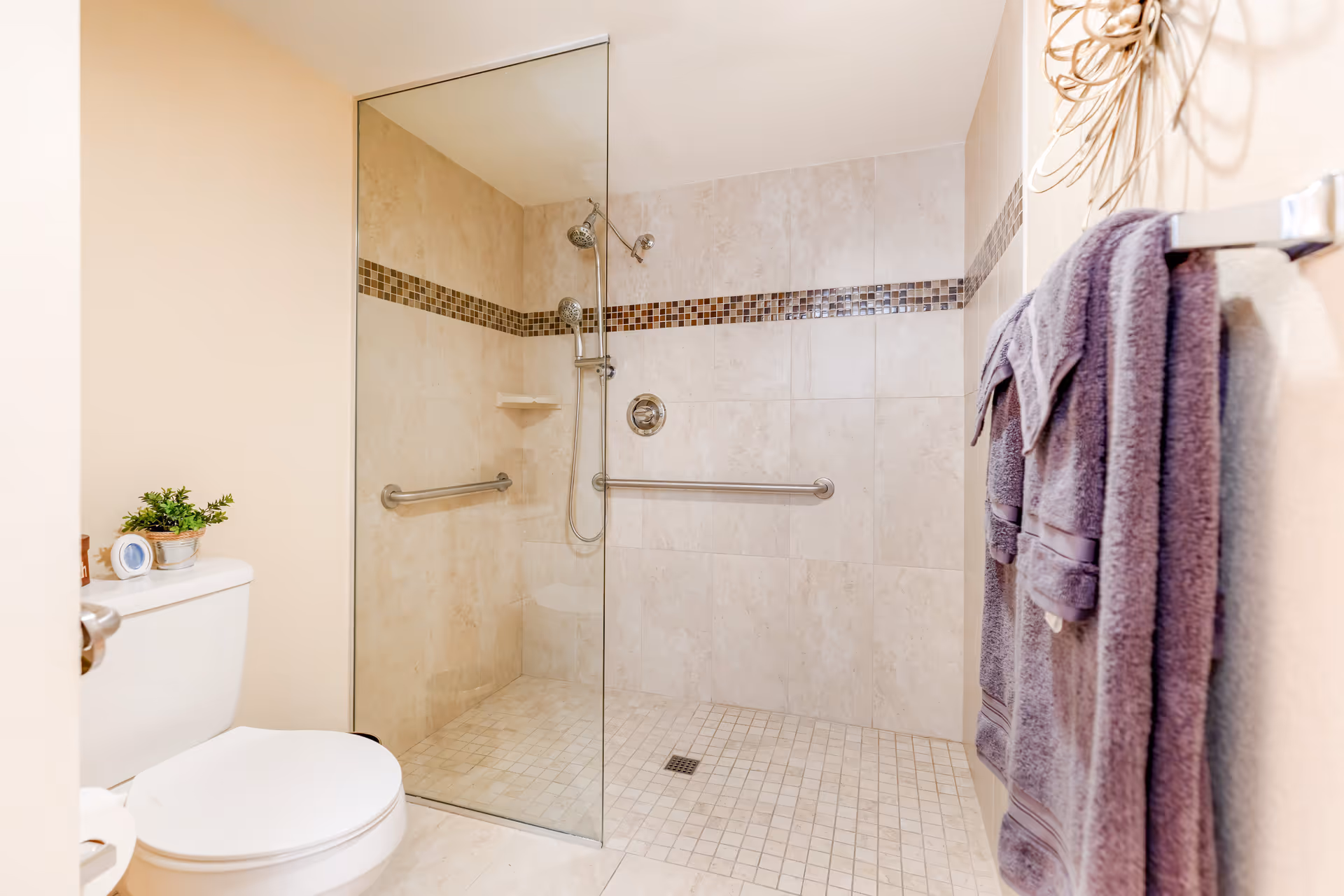 A modern bathroom featuring a walk-in shower with beige tiled walls and floor, a glass partition, and a horizontal mosaic tile accent strip. The shower has two grab bars and a handheld showerhead. To the left, there is a white toilet with a small potted plant and a clock on the tank. On the right wall, two purple towels hang on a towel rack.