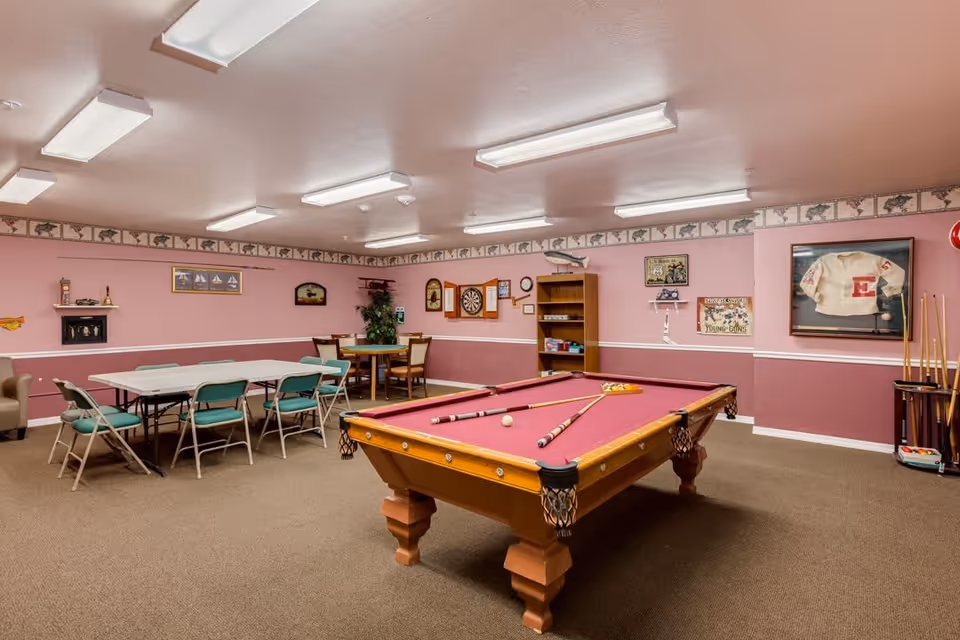 A recreational room with a pool table in the center featuring a red felt surface and pool cues resting on it. To the left, there are two folding tables with green folding chairs around them. The walls are painted pink with a decorative border near the ceiling. The room also contains a dartboard, a bookshelf, framed pictures, and a display case with a sports jersey. The floor is carpeted and the ceiling has multiple fluorescent lights.