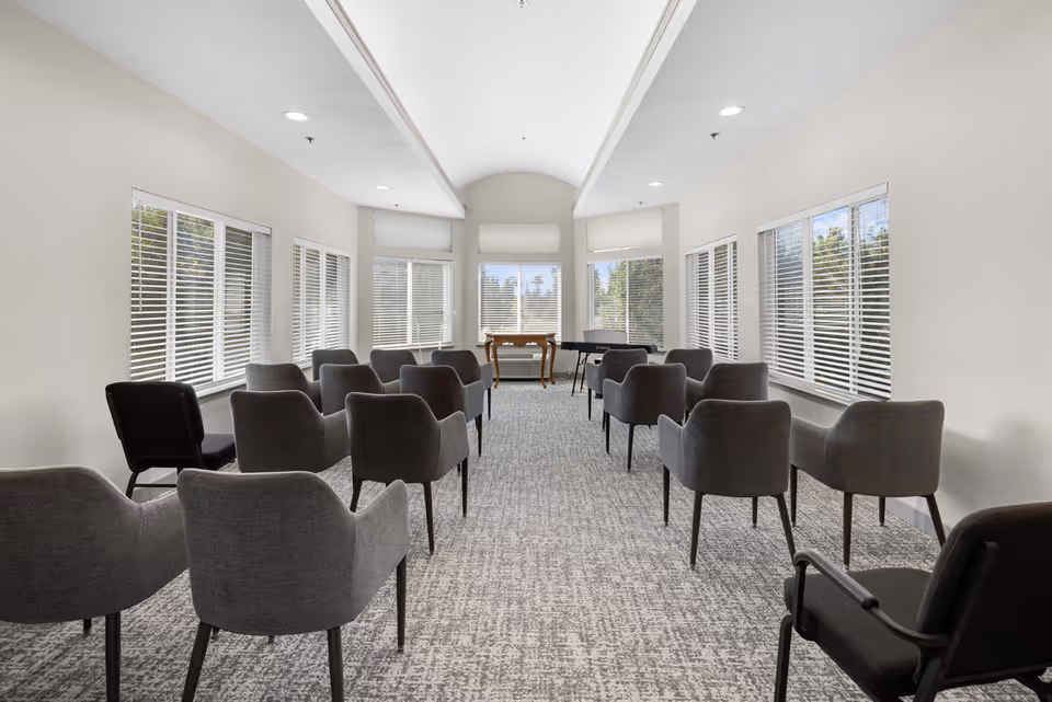 A bright, spacious room with multiple rows of gray upholstered chairs arranged facing a small wooden table and a black piano near large windows with white blinds. The room has light-colored walls, a patterned carpet, and a high ceiling with recessed lighting.