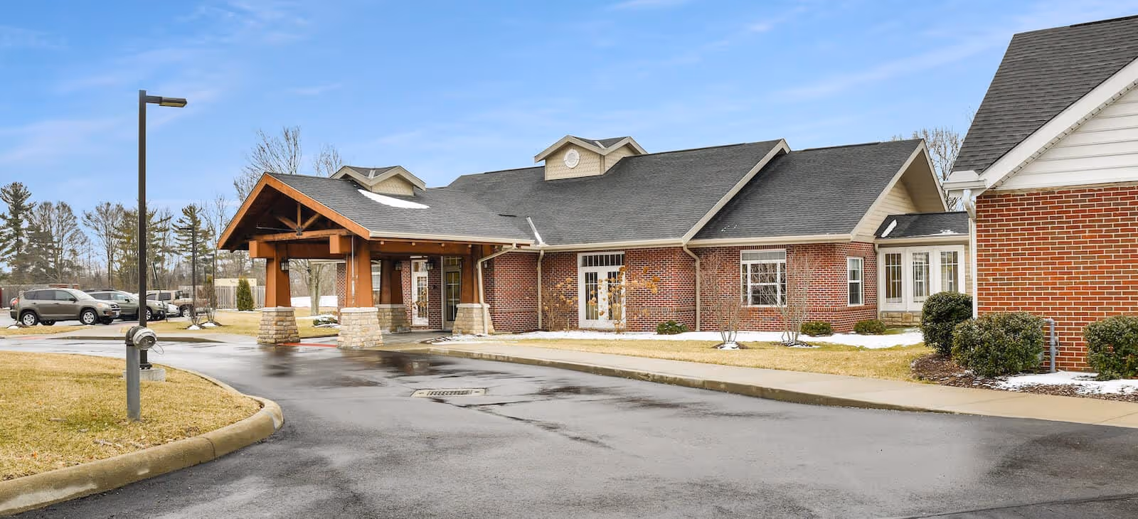 Brick-front senior living facility entrance with a covered porte-cochère, driveway, parked cars, and surrounding lawn.