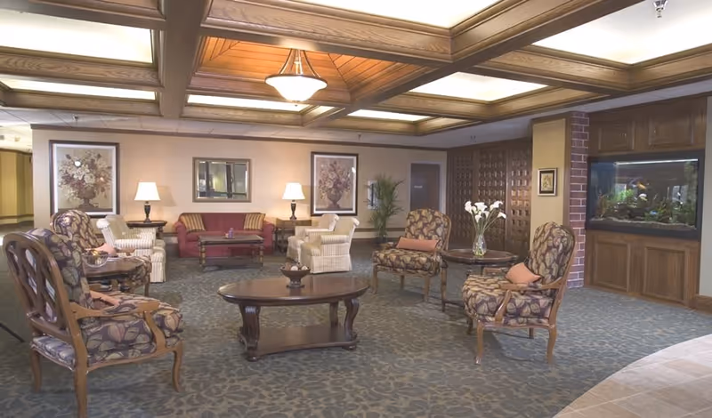 A spacious and well-lit living room area in a senior living facility featuring multiple upholstered chairs with floral patterns, a red sofa, two white armchairs, wooden coffee tables, decorative lamps, framed floral artwork on the walls, a large built-in aquarium, and a ceiling with wooden beams and recessed lighting.