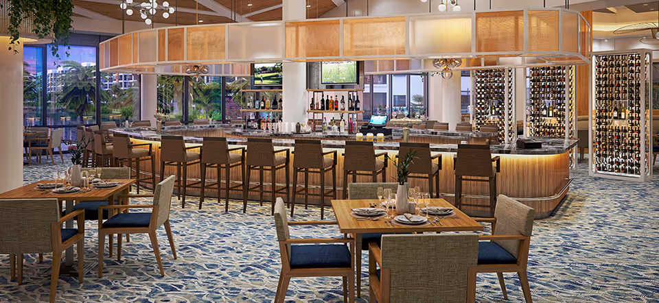 A modern dining area featuring a large bar with high chairs, tables set with plates, glasses, and cutlery, and a wine display on the right side. The space has large windows showing an outdoor view with palm trees and buildings, and the floor is covered with a patterned carpet.