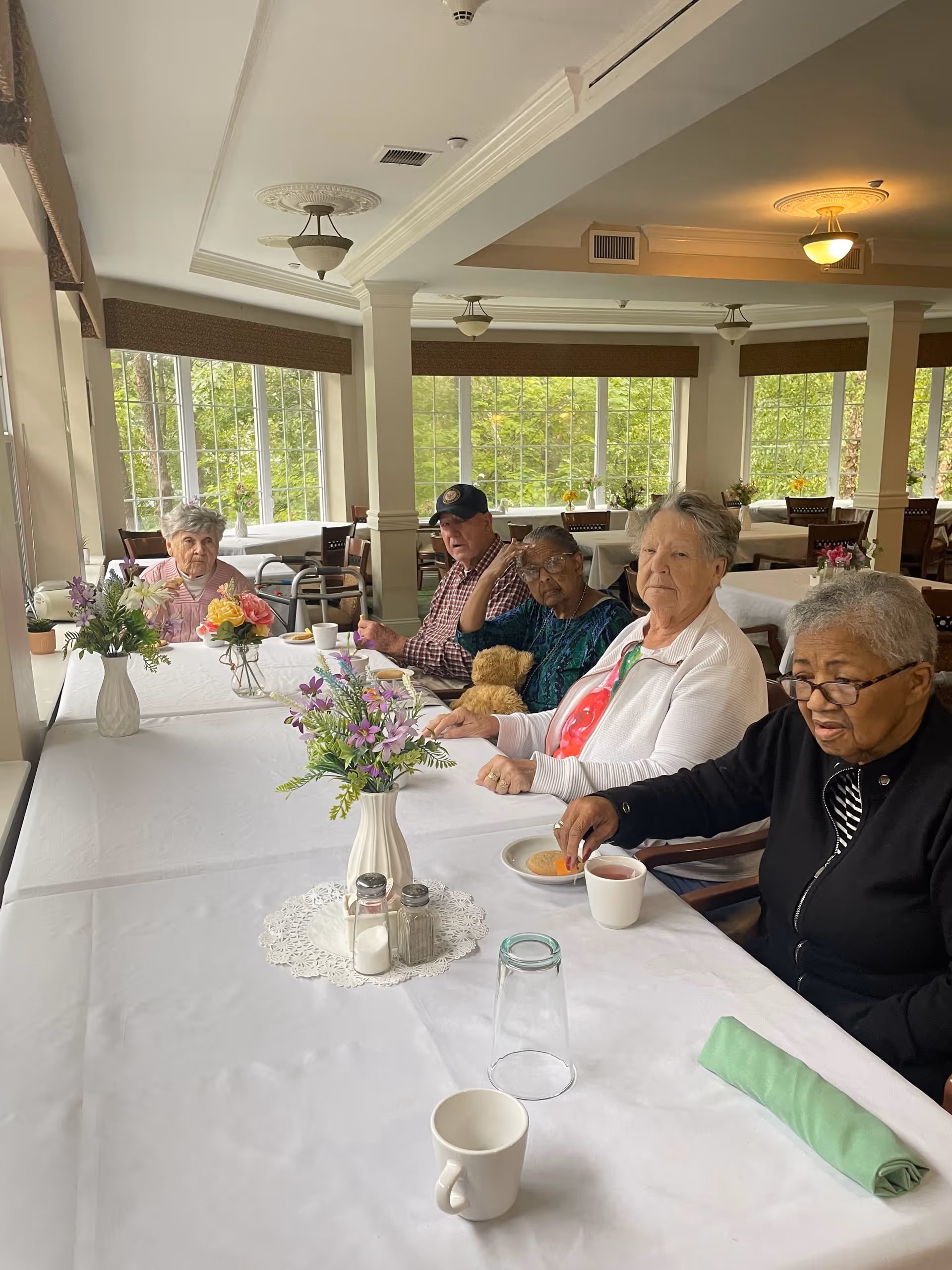 Five elderly individuals sitting around a long dining table in a bright room with large windows. The table is covered with a white tablecloth and decorated with vases of flowers, salt and pepper shakers, and cups. The room has cream-colored walls and ceiling lights, with greenery visible outside the windows.