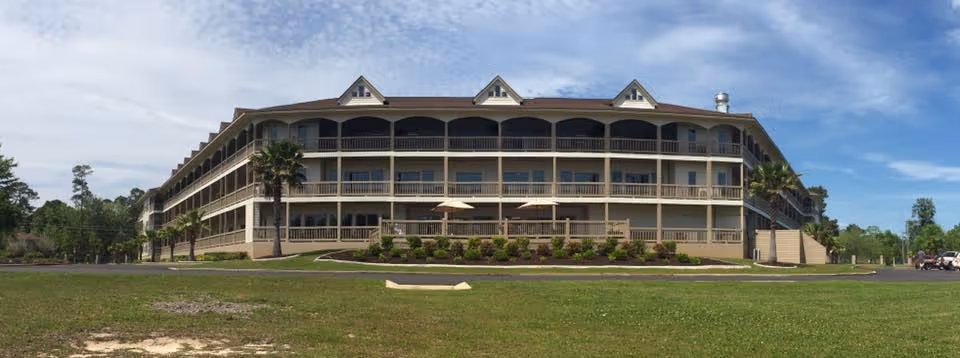 Wide exterior view of a three-story senior living facility building with multiple balconies and a large grassy area in front under a partly cloudy sky.