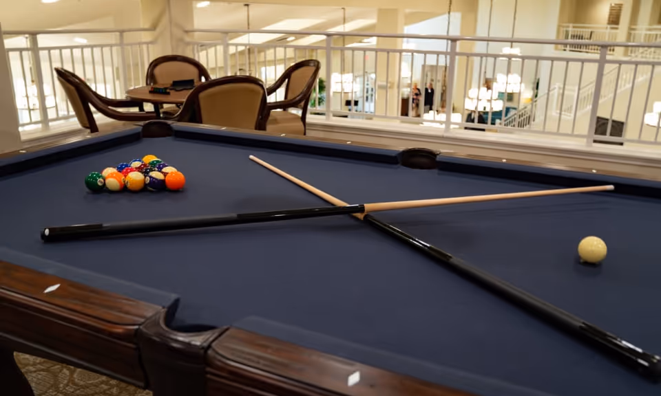 A pool table with a blue felt surface, two crossed pool cues, a rack of billiard balls, and a cue ball. In the background, there are several chairs and a table, with a railing and a well-lit interior space visible beyond.