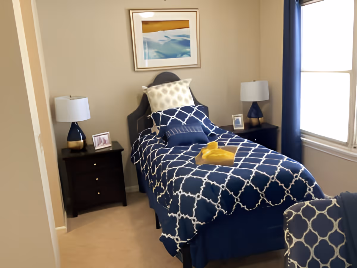A neatly made single bed with a navy blue and white patterned comforter and several pillows in a small bedroom. There are two dark wooden nightstands on either side of the bed, each with a lamp and a small framed picture. A window with blue curtains allows natural light into the room, and a framed abstract painting hangs on the wall above the bed.
