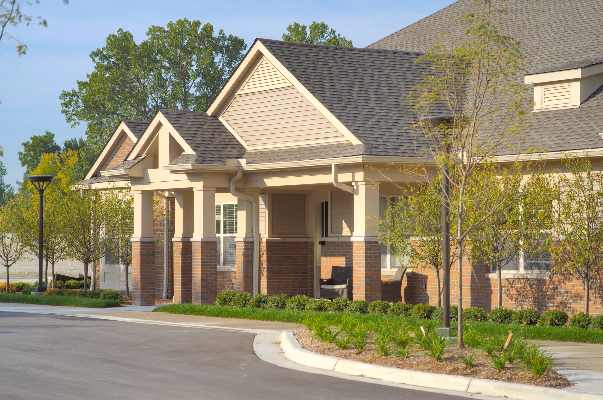 Covered entrance of a single-story brick and beige-siding building with a landscaped driveway and small trees.