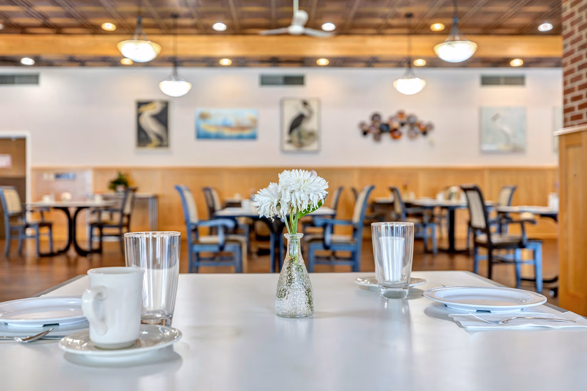 A dining table set with a white cup and saucer, two empty glasses, and two white plates with silverware. A small glass vase with white flowers is centered on the table. In the background, there are multiple dining tables and chairs, wall art, and hanging light fixtures in a well-lit dining room.