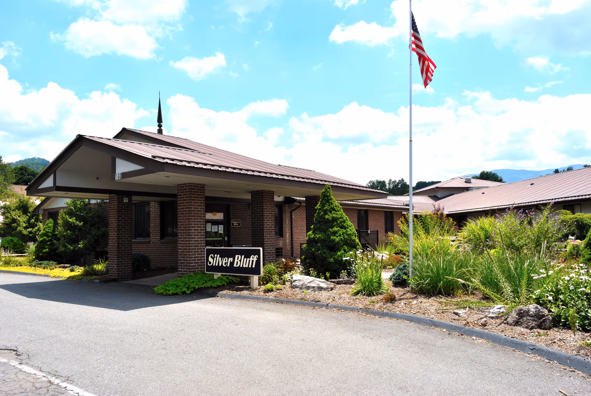 Exterior view of the Silver Bluff senior living facility showing a brick building with a covered entrance, surrounded by landscaped greenery and an American flag on a flagpole.