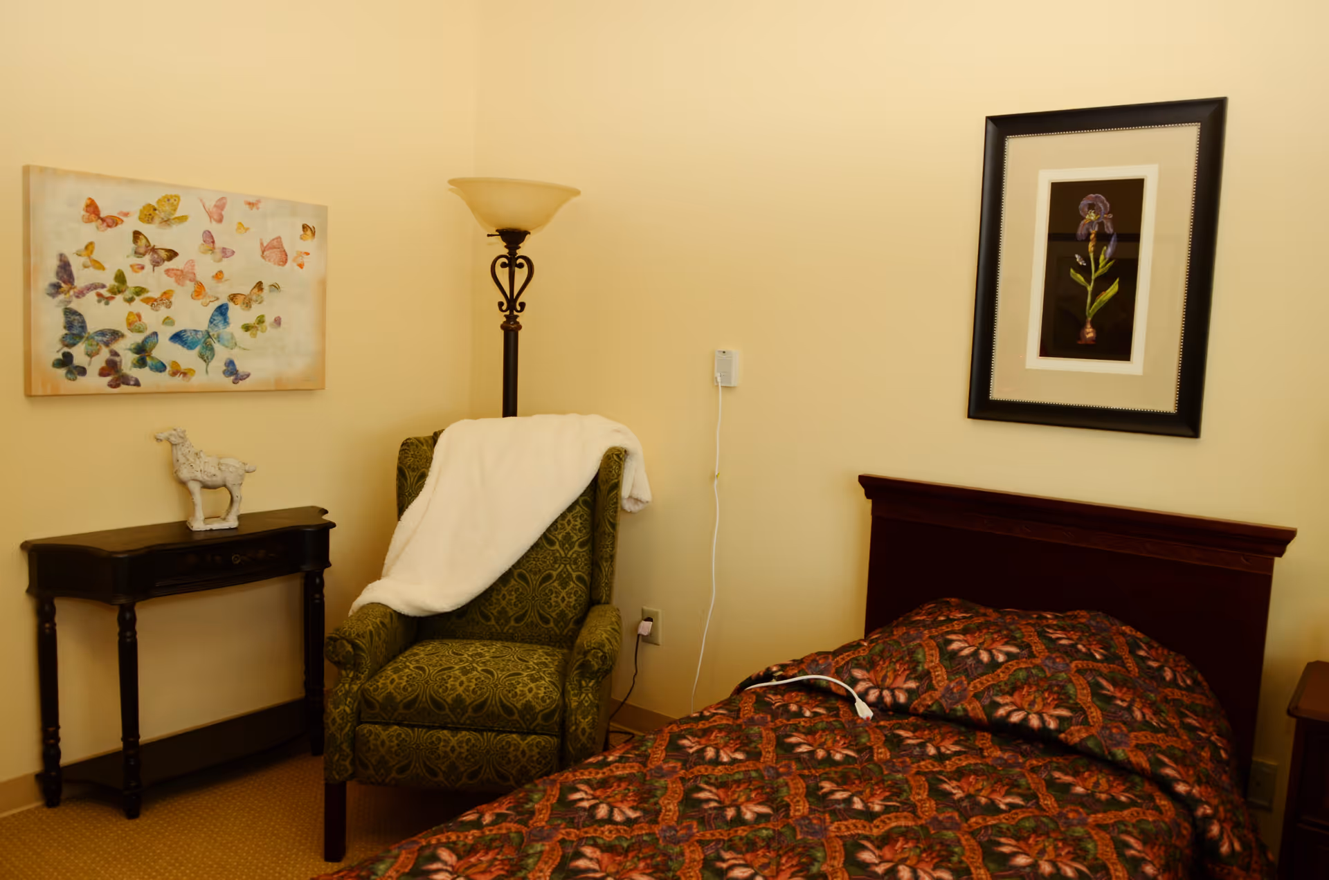 Cozy single-occupancy bedroom with a bed, patterned armchair draped with a blanket, side table, floor lamp, and framed wall art.