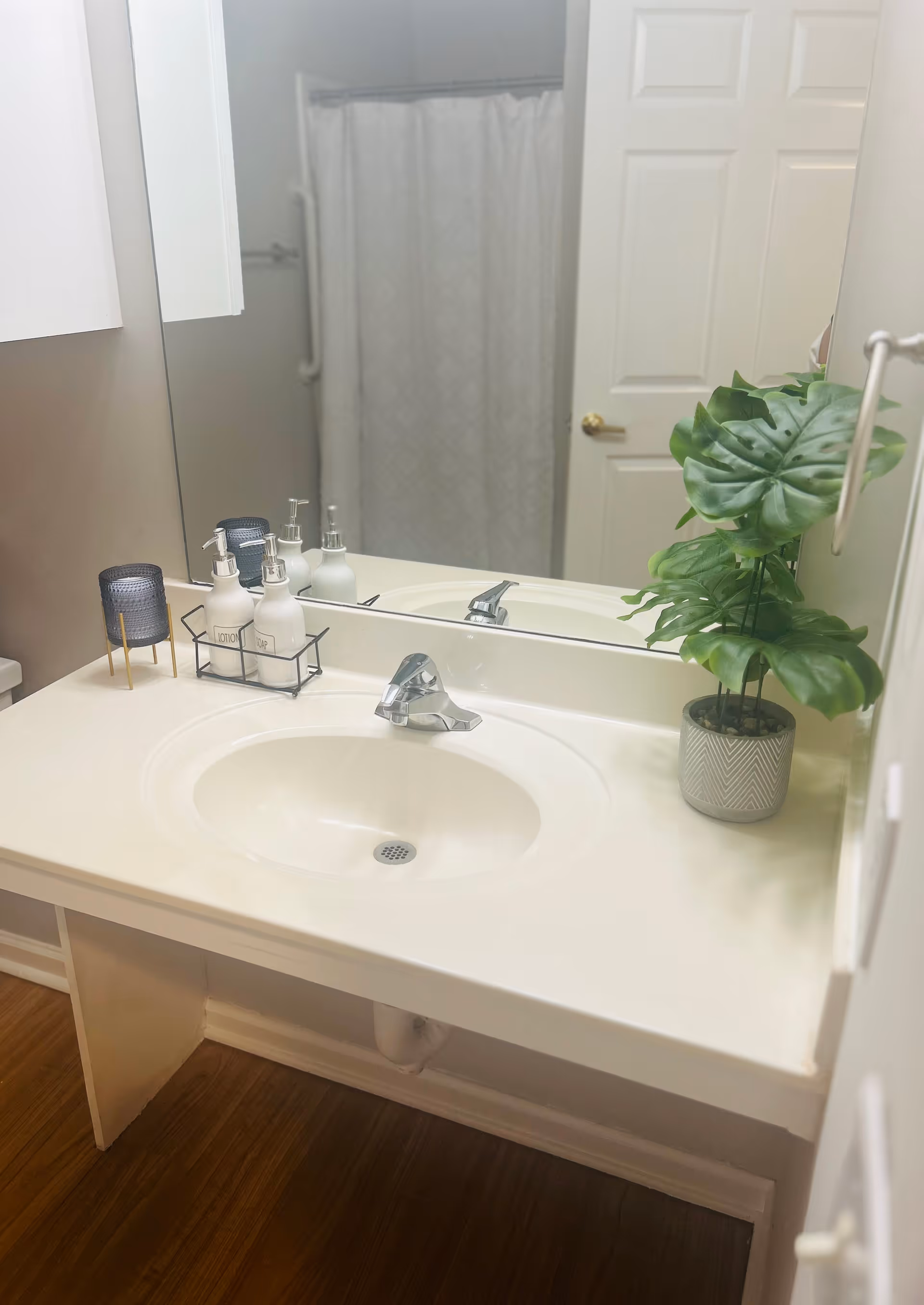 A clean bathroom sink with a silver faucet, a large mirror above it, and a white countertop. On the countertop, there are three pump bottles labeled lotion, soap, and shampoo in a black wire holder, a small dark blue candle holder, and a potted green plant. In the background, a white door with a gold handle and a shower curtain are visible.