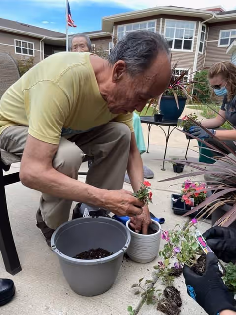 An elderly man wearing a yellow shirt is planting flowers in a white pot outdoors on a concrete patio. Nearby, a woman wearing a mask and gloves is also engaged in planting activities. There are various potted plants and gardening supplies around them, with a building and an American flag visible in the background.