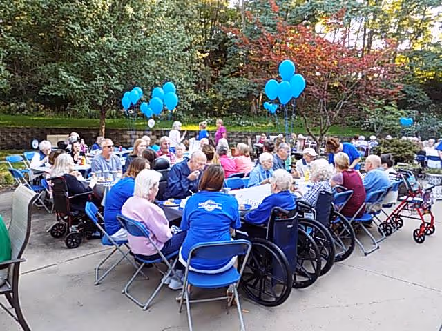 A large group of elderly people seated outdoors around several tables, some in wheelchairs and others in chairs, enjoying a social gathering with blue balloons tied to the tables. The setting is a paved patio area surrounded by trees and greenery.