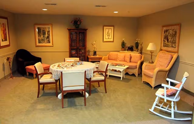 A cozy living room with a round table covered with a lace tablecloth surrounded by four chairs. There is a peach-colored sofa, two matching armchairs, a white rocking chair with a cushion, and a dark armchair with an orange cushion. The walls are decorated with framed artwork and a wooden cabinet with flowers on top.