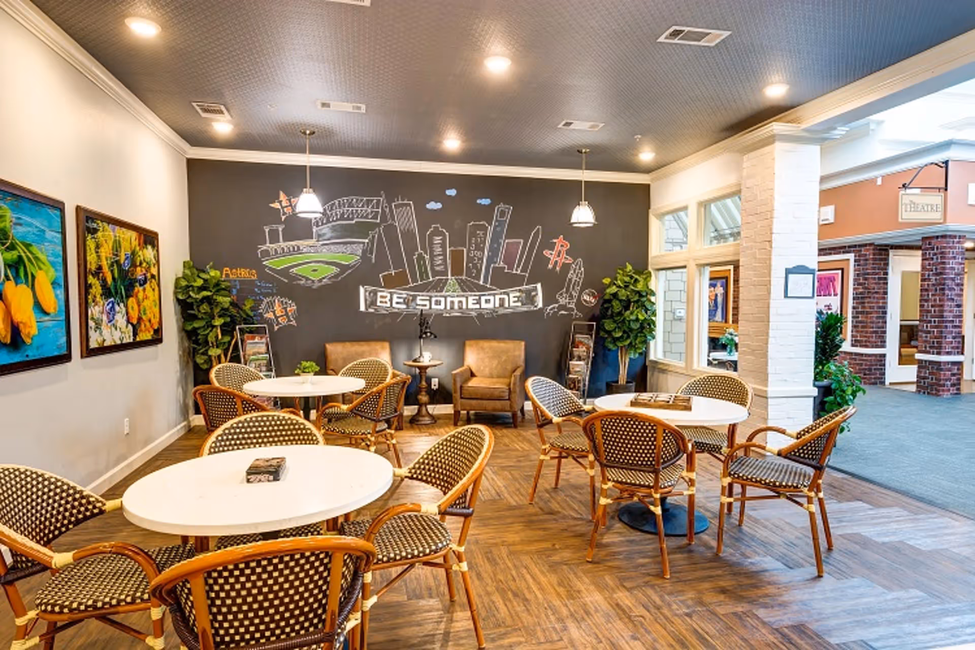 A bright and inviting common area with round white tables and wicker chairs arranged on a wood-patterned floor. The back wall features a large blackboard with colorful chalk drawings of a city skyline, a baseball field, and the words 'BE SOMEONE'. Two brown armchairs and a small table are positioned against the blackboard wall. The room is well-lit with ceiling lights and pendant lamps, and there are framed floral paintings on the left wall. Large windows and an open doorway lead to an adjacent hallway with brick and white pillars.