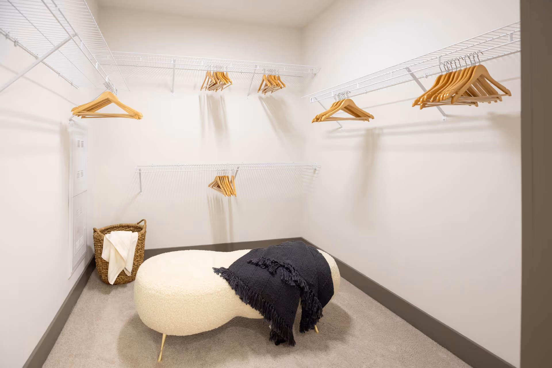 A spacious walk-in closet with white wire shelving and multiple wooden hangers. There is a woven basket with a white cloth inside on the floor and a cream-colored cushioned bench with a black throw blanket draped over it. The walls are painted white and the floor is carpeted in a light beige color.