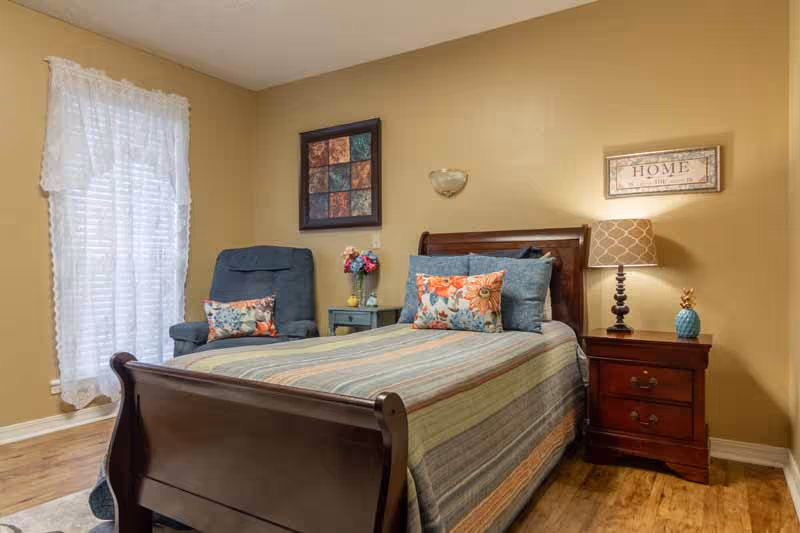 A cozy bedroom with a wooden bed frame, a striped bedspread, and decorative pillows. There is a blue armchair with a floral pillow next to a small side table holding a vase with flowers. The room has beige walls, a window with white lace curtains, a framed artwork above the armchair, and a framed sign that reads 'HOME' above a wooden nightstand with a lamp and a small pineapple decoration.