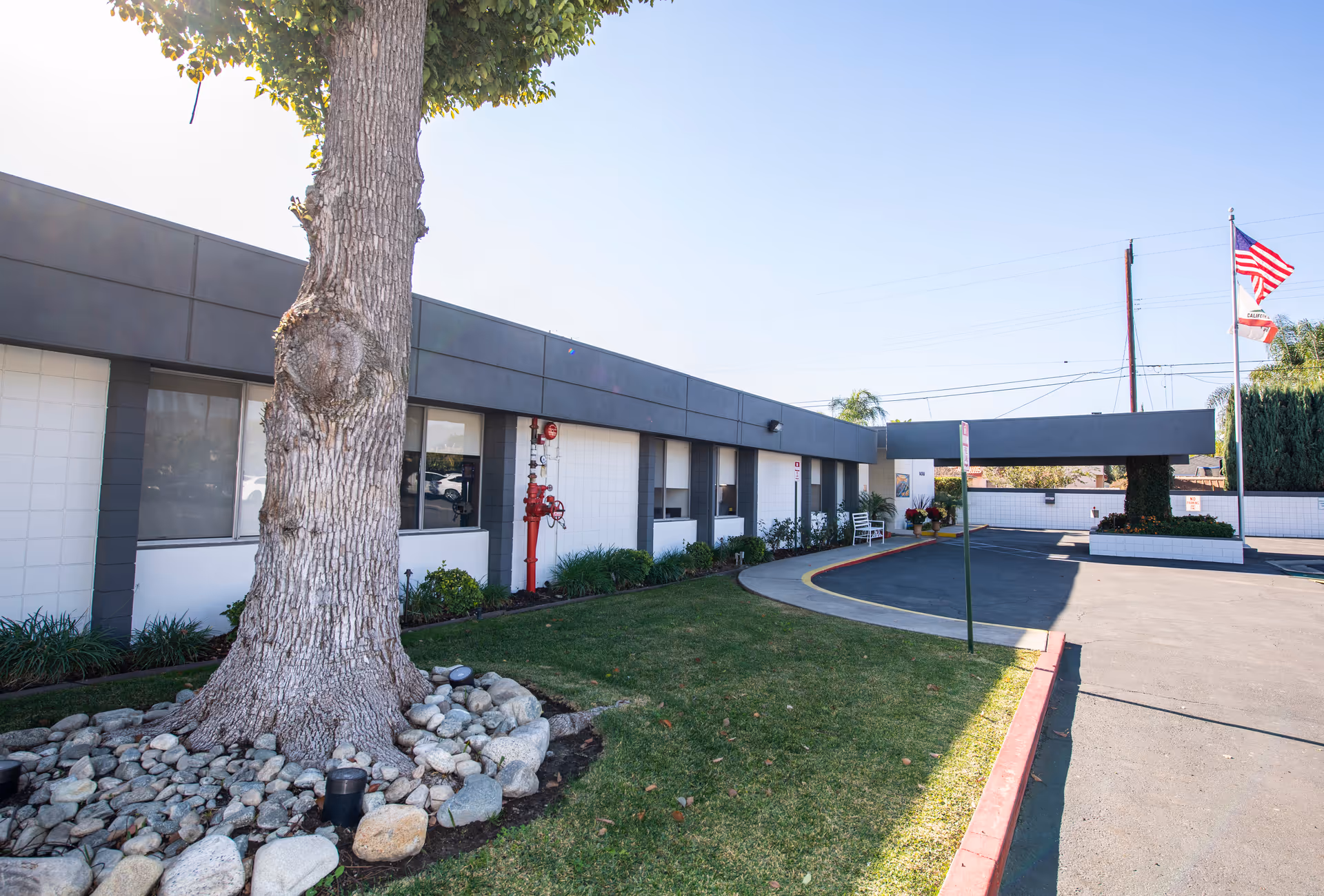 Exterior view of Sierra View Care Center showing a single-story building with white walls and dark trim, a large tree surrounded by rocks in the foreground, a curved driveway, and two flagpoles with the American and California state flags.