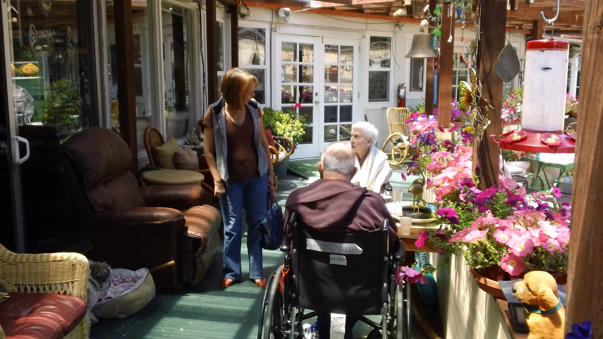 A covered outdoor patio area at a senior living facility with two elderly people seated and a woman standing nearby. The space is decorated with vibrant pink flowers, wicker and leather chairs, and a bird feeder hanging from a wooden beam. One elderly person is in a wheelchair facing away from the camera, while the other is seated and wrapped in a white blanket.