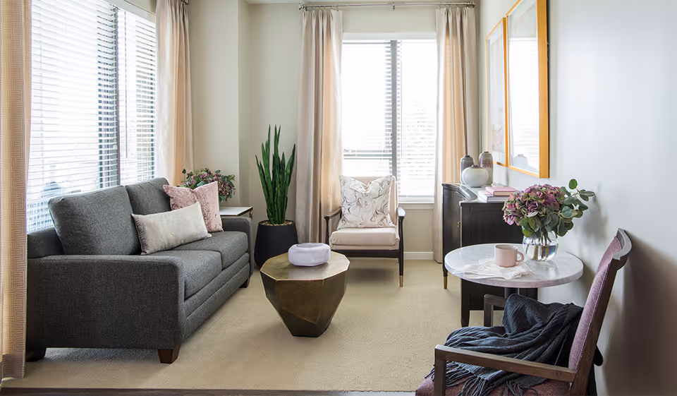 A cozy living room with a gray sofa adorned with pillows, a beige armchair, a geometric coffee table, and a small round table with a vase of flowers and a pink mug. The room has large windows with beige curtains, a potted plant, and framed artwork on the wall.