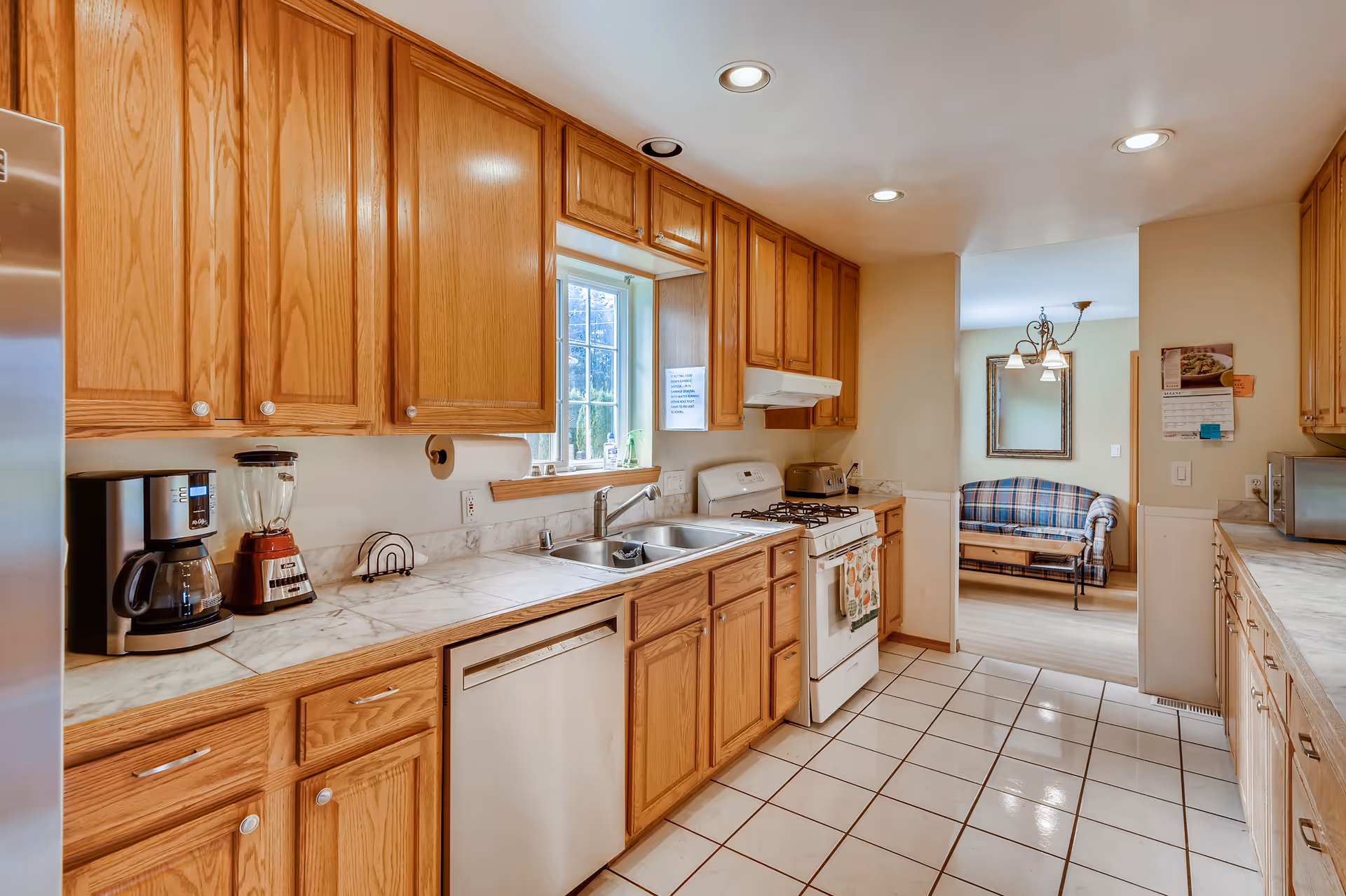 A bright kitchen with wooden cabinets, white tiled floor, and marble countertops. The kitchen features a coffee maker, blender, dishwasher, double sink, gas stove with oven, and a microwave. A window above the sink lets in natural light. In the background, a living area with a plaid sofa and a wall mirror is visible.