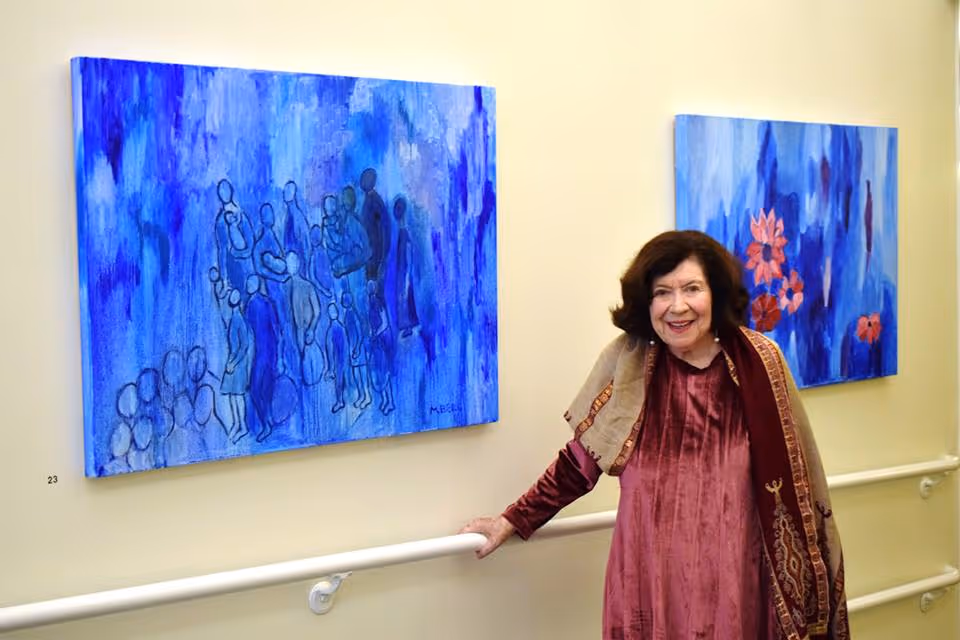 An elderly woman wearing a maroon dress and a patterned shawl stands smiling in a hallway next to two blue-themed paintings on the wall. She is holding onto a white handrail attached to the wall.