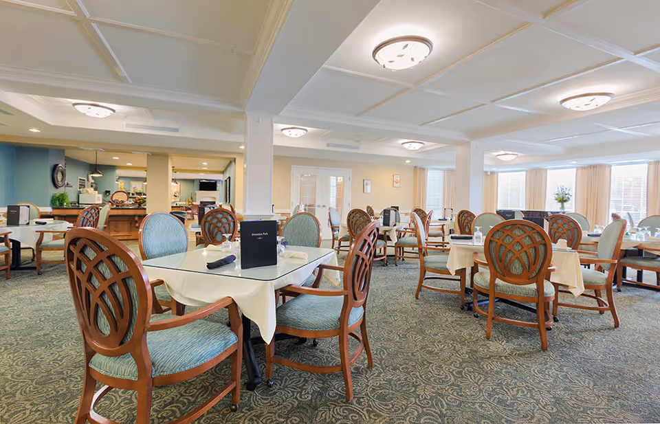 A spacious and well-lit dining room with multiple tables covered in white tablecloths and set with glassware and napkins. The chairs have wooden frames with intricate back designs and light blue upholstery. The room features patterned carpet, ceiling lights, large windows with beige curtains, and a buffet area in the background.