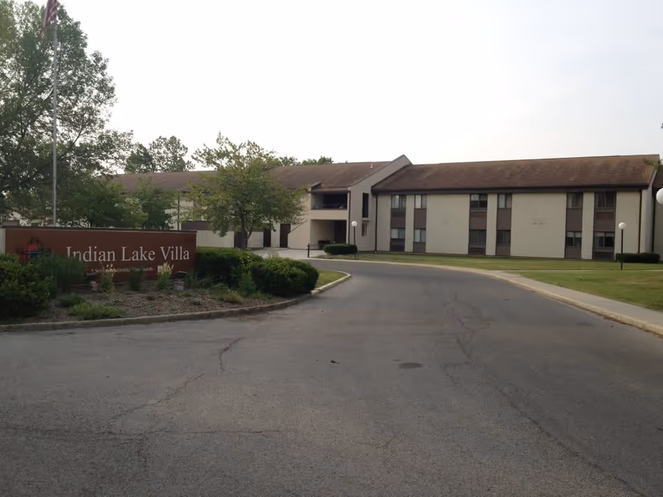 Exterior view of Indian Lake Villa, a senior living community building with a two-story structure, surrounded by trees and greenery, with a paved driveway leading to the entrance and a sign displaying the facility name.