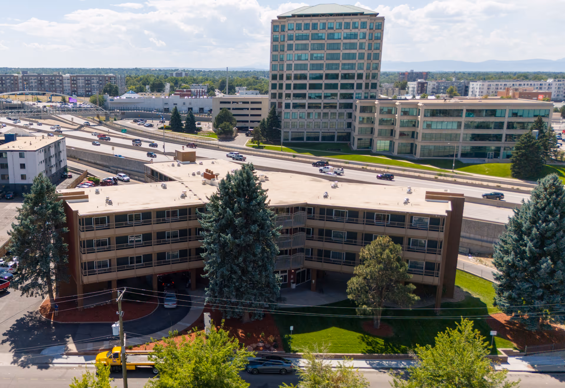 Aerial view of a mid-rise rehabilitation facility building with surrounding trees, parking area, and a highway with office buildings in the background.
