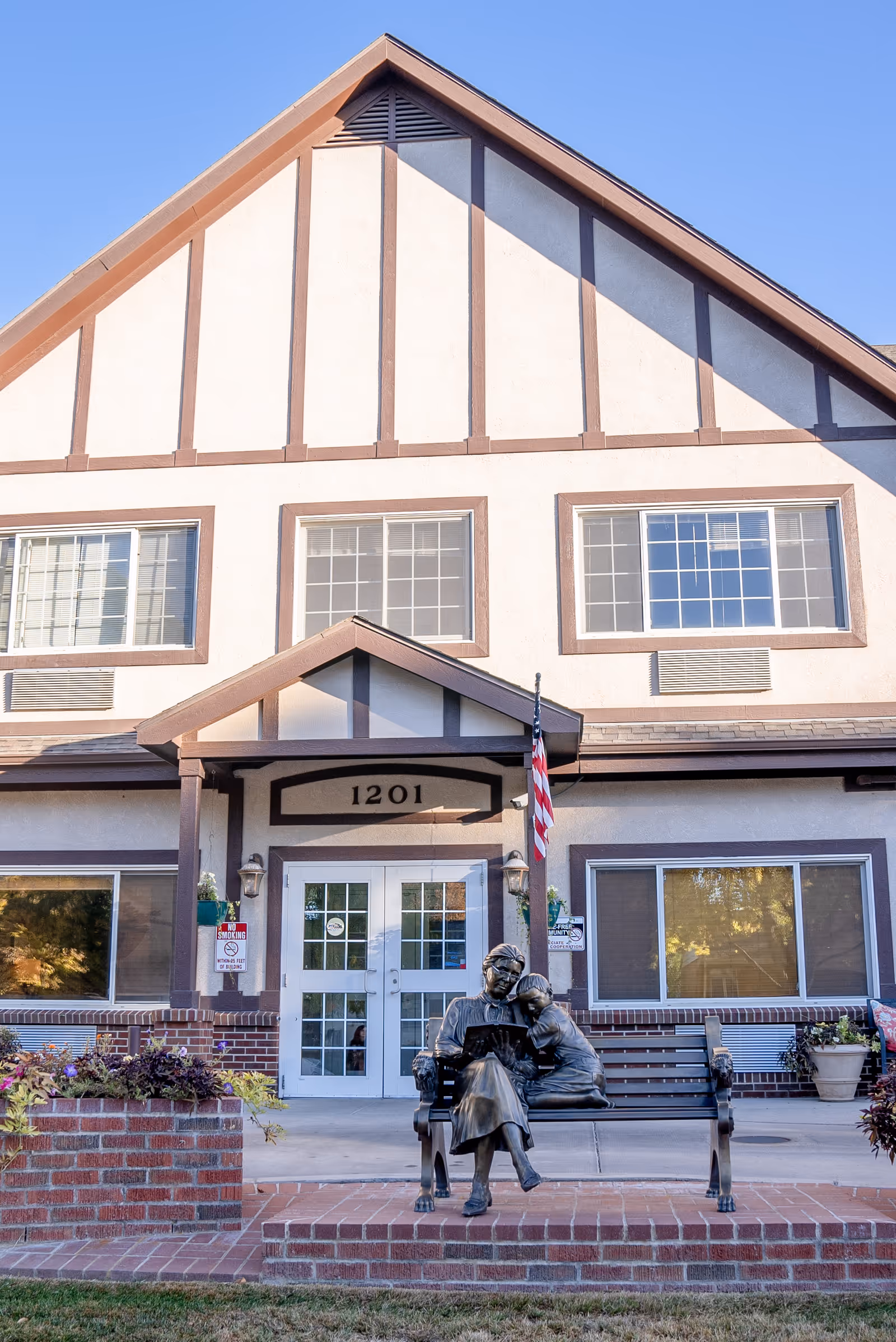 Front exterior view of Chateau At Sharmar Assisted Living building with a peaked roof, large windows, and a double glass door entrance. In front of the entrance is a bench with a bronze statue of an elderly woman reading a book to a child. There are flower beds and an American flag near the entrance.
