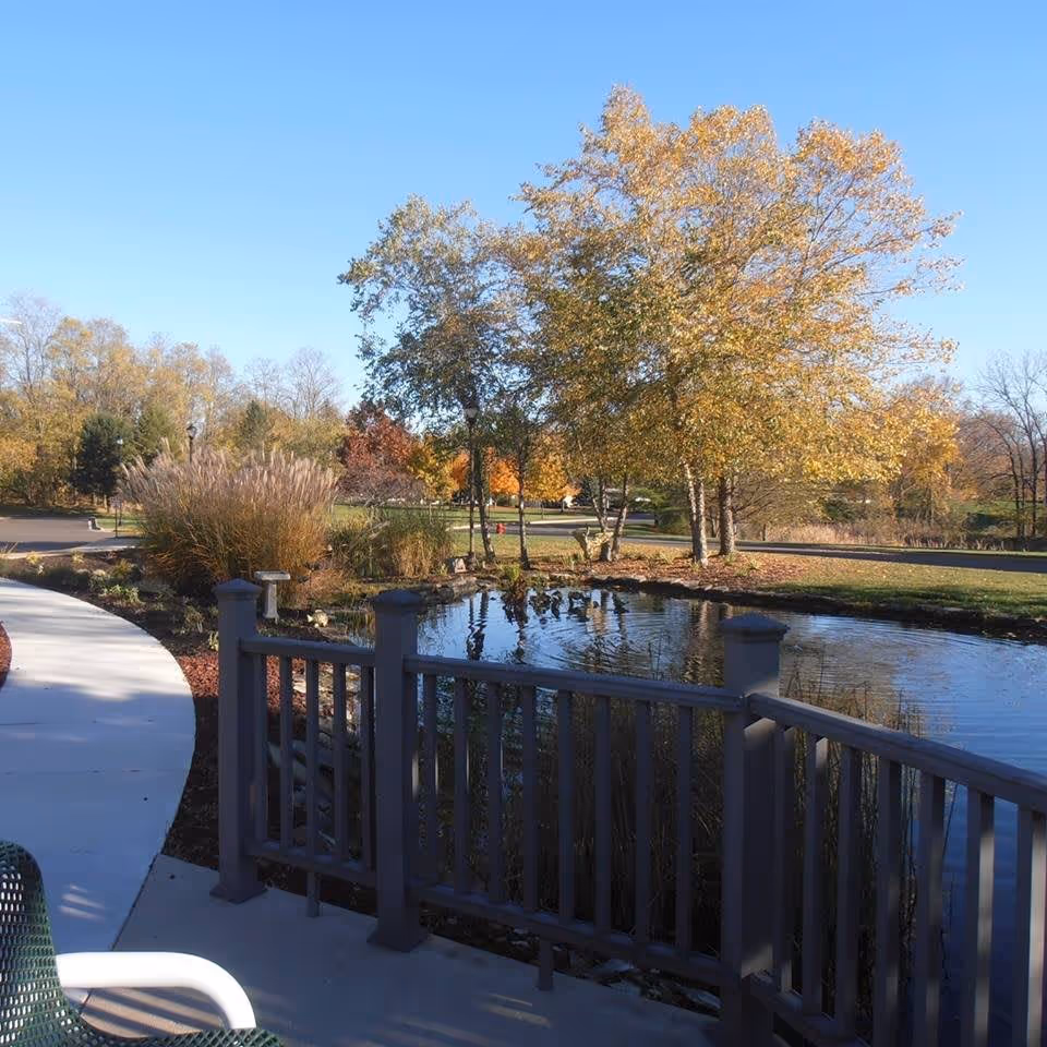 Outdoor scene at Marsh Pointe featuring a small pond with a wooden railing in the foreground, surrounded by autumn trees with yellow and orange leaves, a curved concrete walkway, and a clear blue sky.