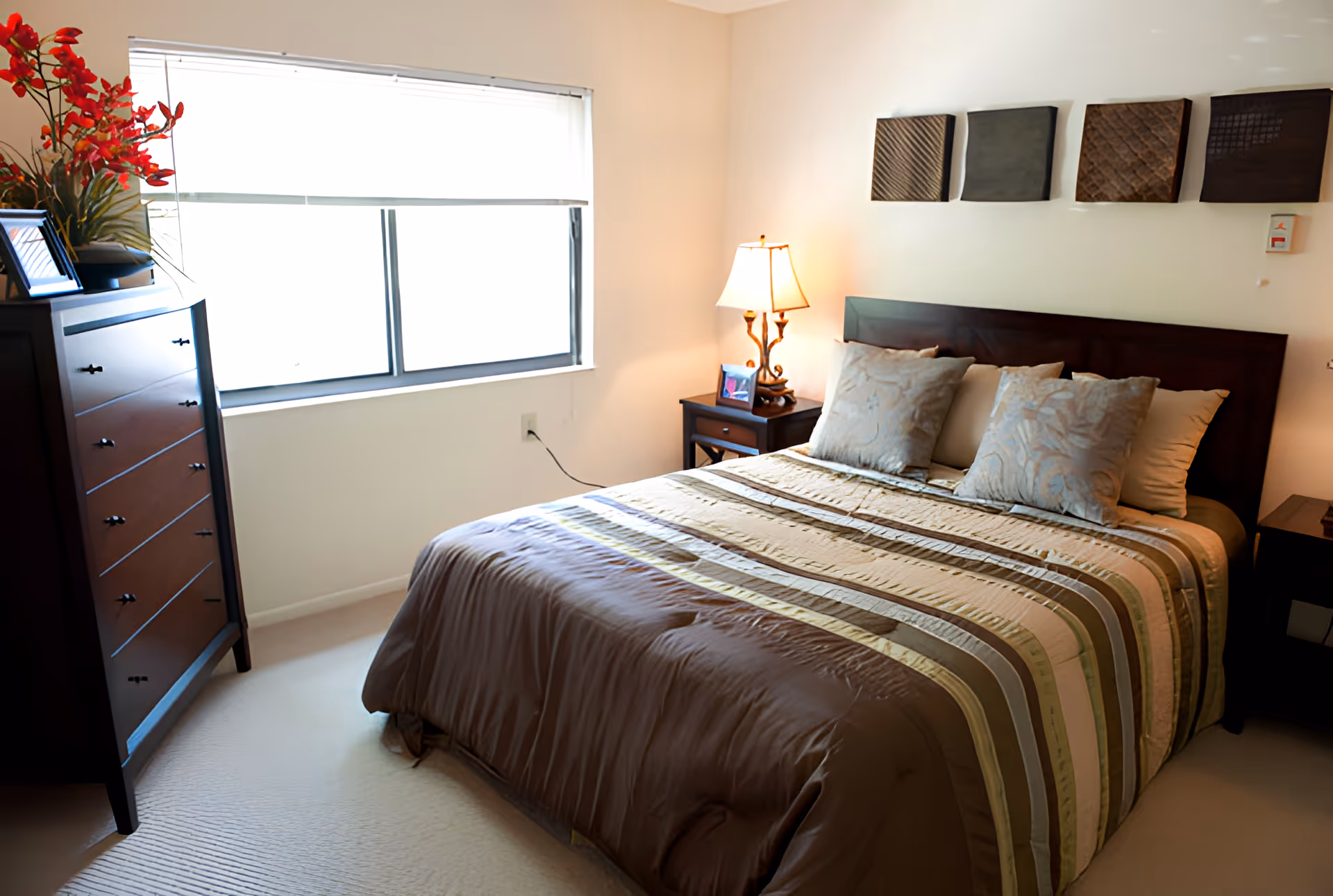 A neatly made bed with a brown and beige striped comforter and four pillows in a bedroom. There is a wooden headboard with four decorative square wall hangings above it. A nightstand with a lamp and a framed photo is beside the bed. Across from the bed is a tall wooden dresser with a vase of red flowers and a framed photo on top. A window with a white blind is on the wall behind the dresser.