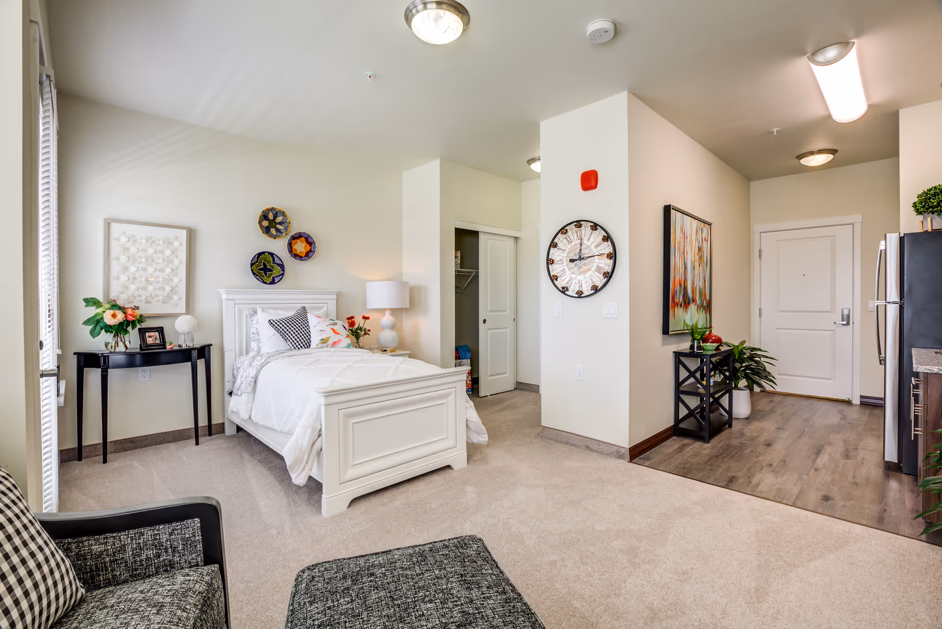 A bright and cozy studio apartment featuring a white bed with decorative pillows and a bedside table with a lamp and flowers. There is a small black table with a plant and picture frame next to the bed. The room has light beige walls and carpet, a large wall clock, and a colorful abstract painting near the entrance door. A small kitchen area with stainless steel appliances and wooden flooring is visible to the right. A comfortable armchair with a checkered pillow is in the foreground.