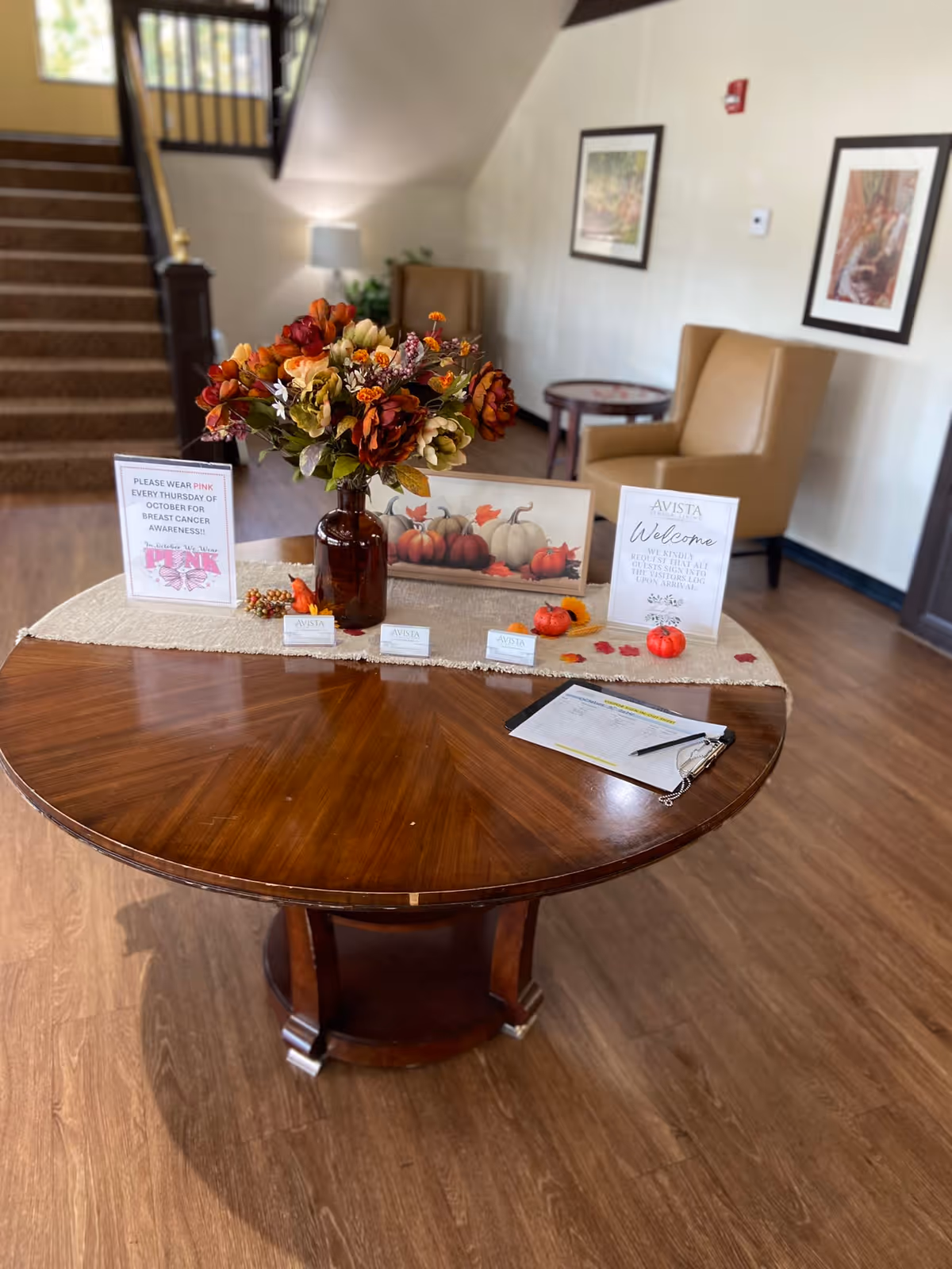 A round wooden table in a senior living facility lobby decorated with a fall-themed floral arrangement in a brown vase, small pumpkins, and informational signs. Behind the table are two beige armchairs, framed artwork on the wall, and a staircase leading upstairs.