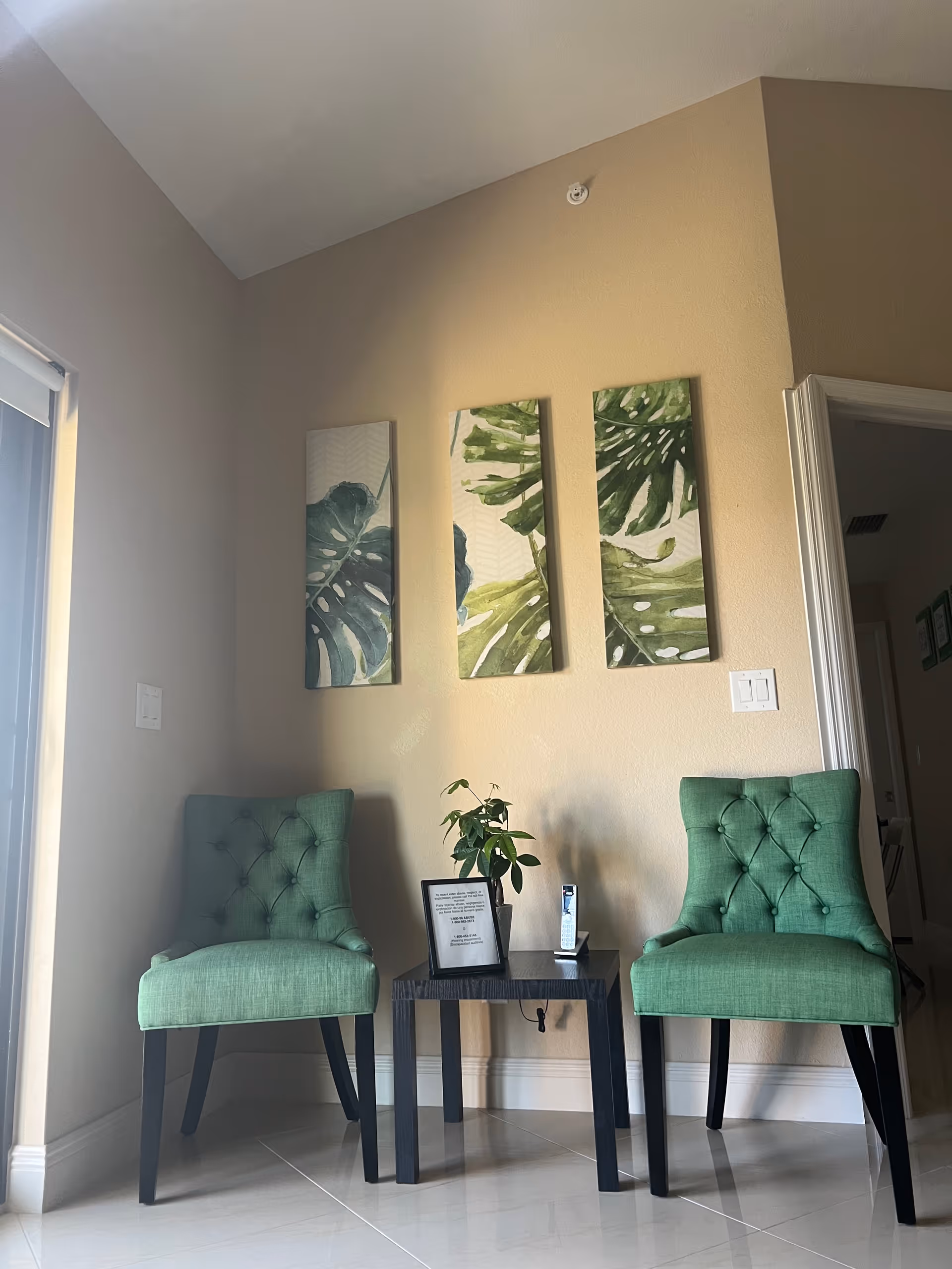 A cozy corner with two green upholstered chairs flanking a small black table. On the table, there is a small potted plant, a framed note, and a cordless phone. Above the chairs, three vertical wall art pieces feature large green leaves. The walls are beige, and the floor is tiled with a glossy finish.