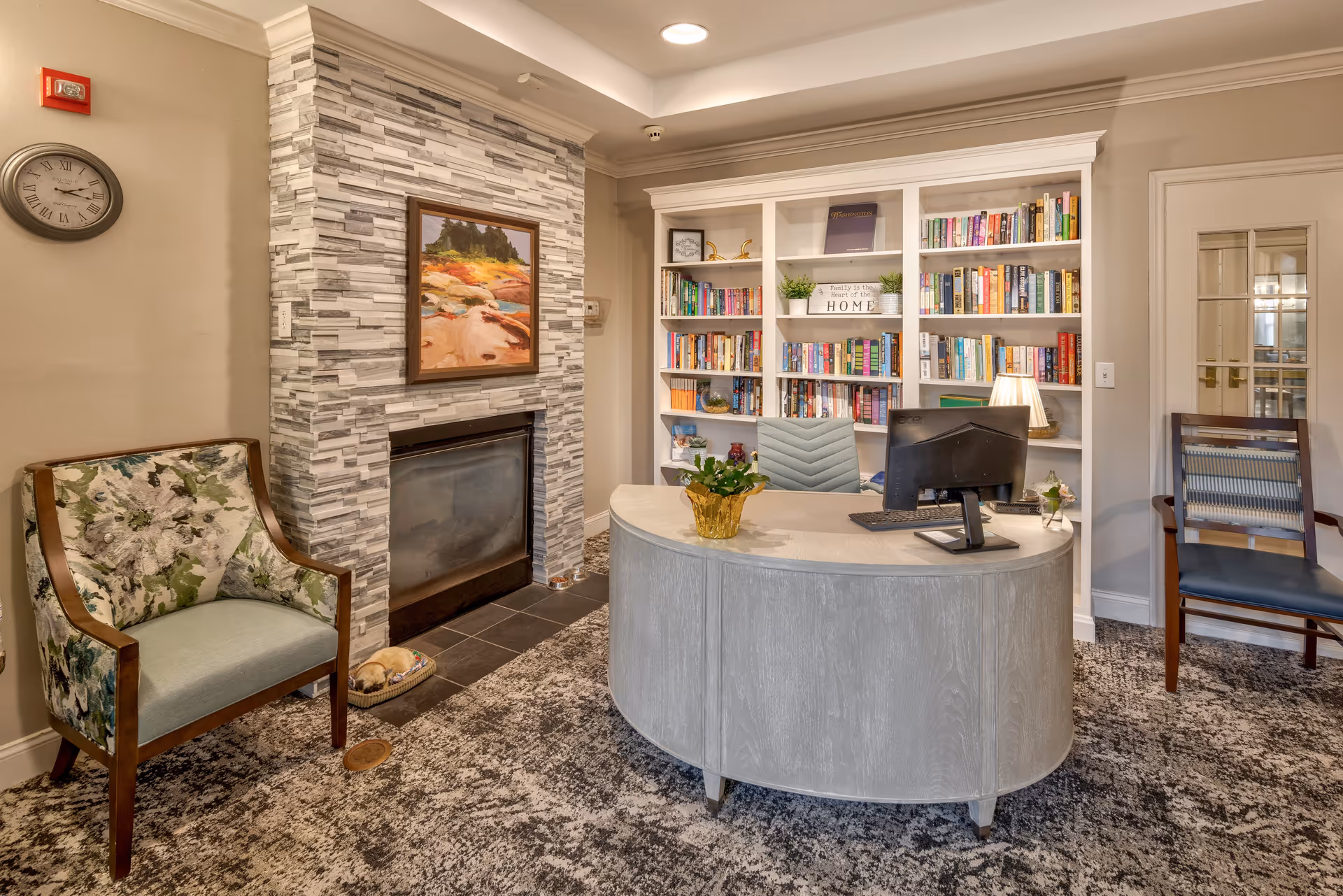 A cozy interior room featuring a curved reception desk with a computer monitor and a small plant on top. Behind the desk is a white built-in bookshelf filled with books and decorative items. To the left, there is a modern fireplace with a stone tile surround and a colorful landscape painting above it. A floral upholstered armchair is positioned near the fireplace, and a clock is mounted on the wall above it. On the right side, there is a wooden chair with a striped cushion and a door with glass panels. The room has a patterned carpet and warm lighting.
