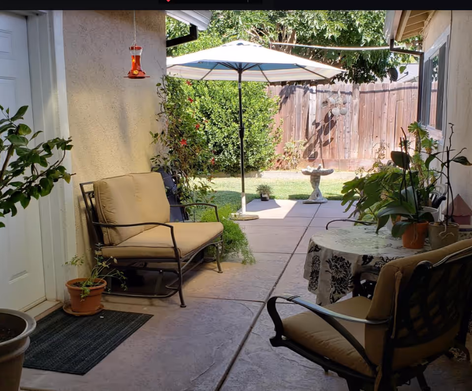 A cozy outdoor patio area with cushioned metal chairs, a small table covered with a floral tablecloth, several potted plants, a hanging red bird feeder, a large umbrella providing shade, and a birdbath near a wooden fence with greenery in the background.