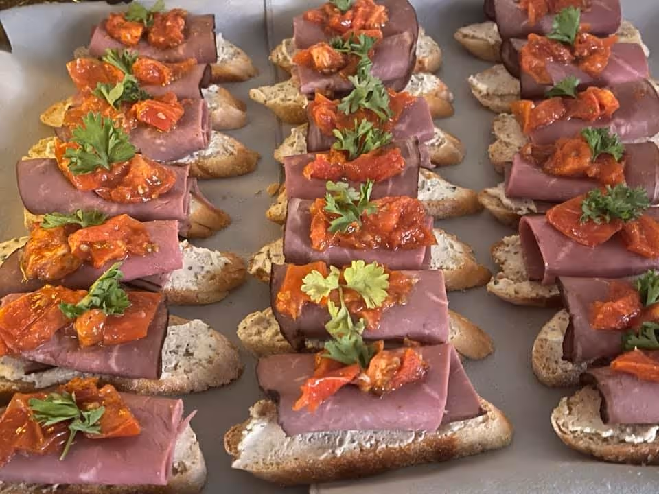 Rows of small open-faced sandwiches with a slice of rolled deli meat, a dollop of red roasted pepper topping, and a sprig of parsley on a slice of bread with a spread.