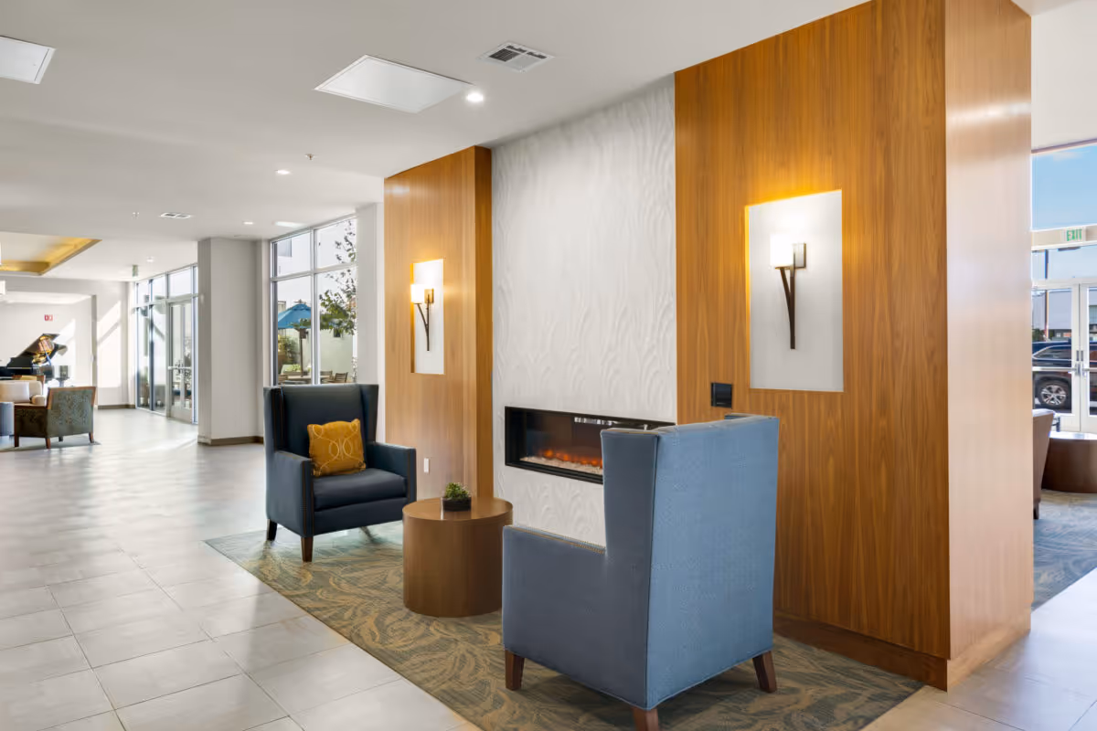 A modern senior living facility lounge area with two blue armchairs facing a built-in electric fireplace set in a white textured wall with wooden panels on either side. A small round wooden table with a small plant is between the chairs. The space has large windows letting in natural light and additional seating and a piano are visible in the background.