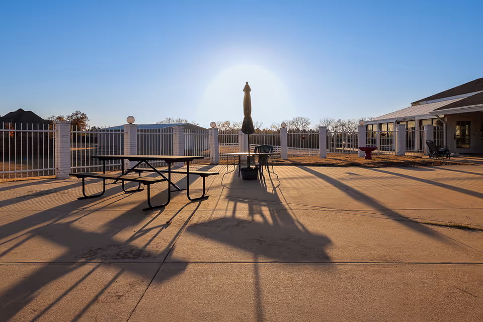 Outdoor patio area at sunset with a picnic table, chairs, and a closed umbrella casting long shadows on the concrete ground, surrounded by a white fence and adjacent to a building with large windows.