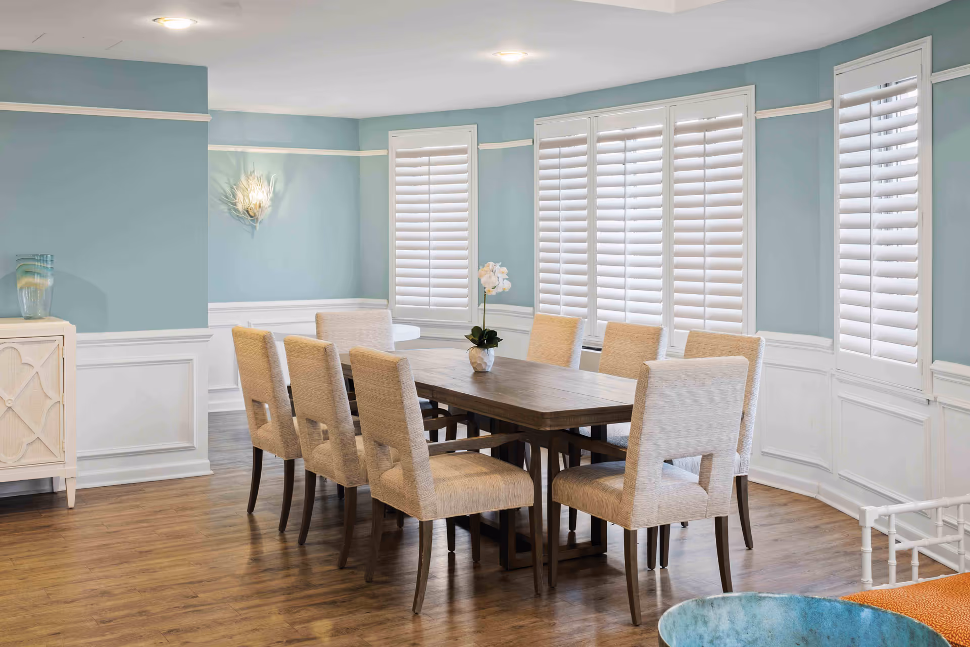 Bright dining room with a wooden table surrounded by eight upholstered chairs and large shuttered windows.