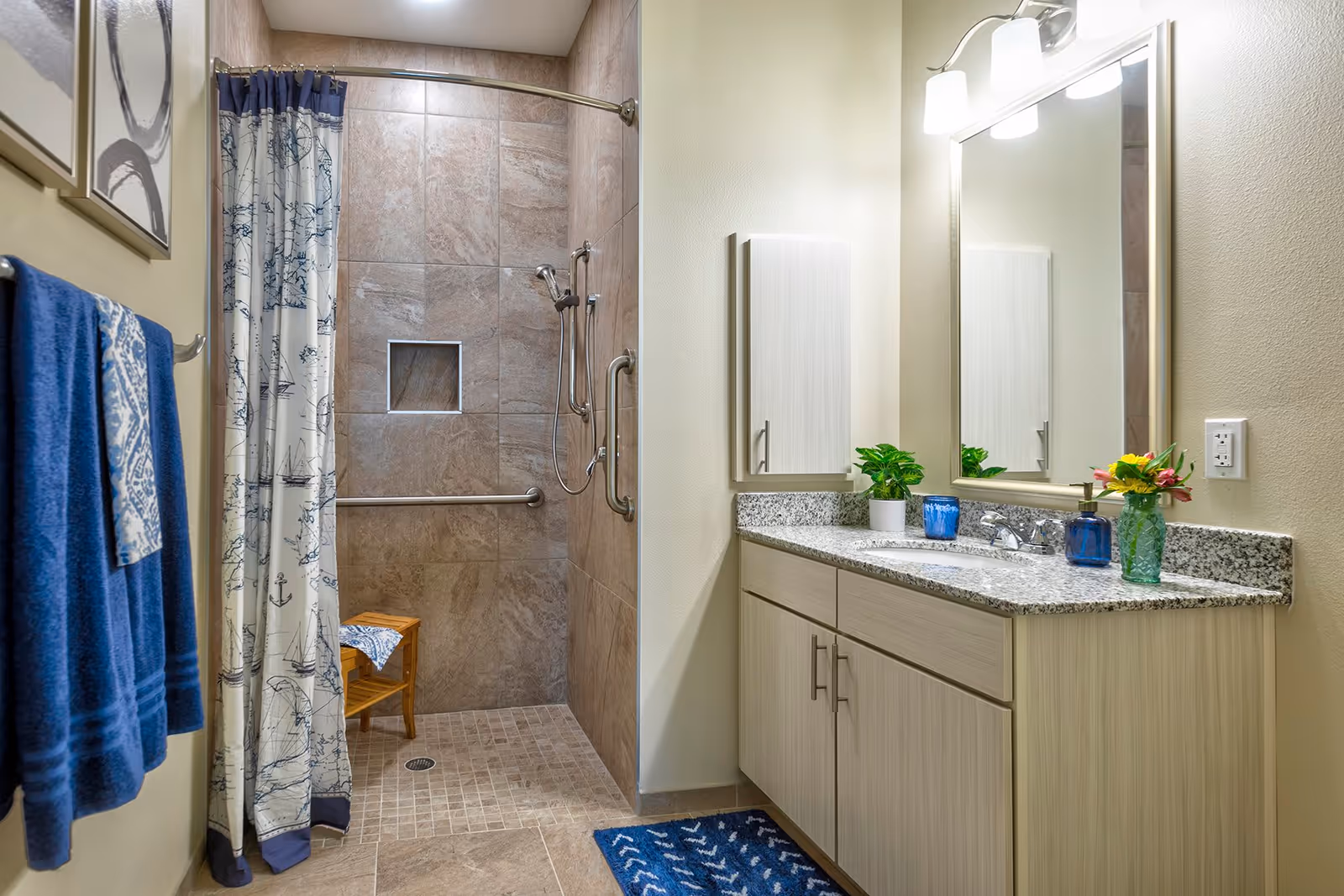 A bathroom with a walk-in tiled shower featuring a handheld showerhead and grab bars. The shower has a nautical-themed curtain and a small wooden stool inside. Next to the shower is a vanity with a granite countertop, a sink, a large mirror, and a cabinet above. The countertop has decorative items including a small plant, a blue candle holder, a soap dispenser, and a vase with flowers. Blue towels hang on a towel rack on the left wall, and a blue patterned rug is on the floor in front of the vanity.
