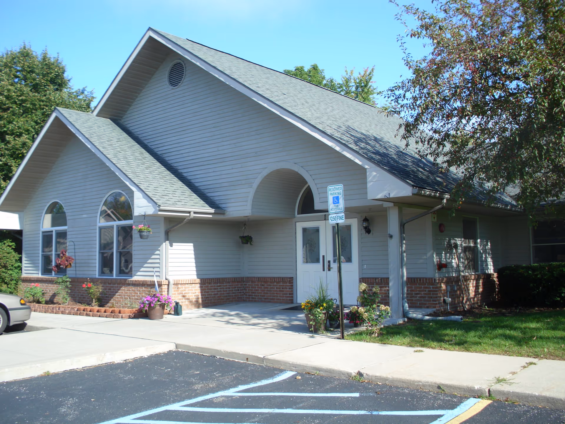 Exterior view of a single-story building with light gray siding and brick accents, featuring a peaked roof and arched windows. There is a concrete sidewalk leading to double doors under a covered entrance. A reserved parking sign for handicapped parking is visible near the entrance, along with some potted plants and landscaping. Trees and greenery surround the building under a clear blue sky.