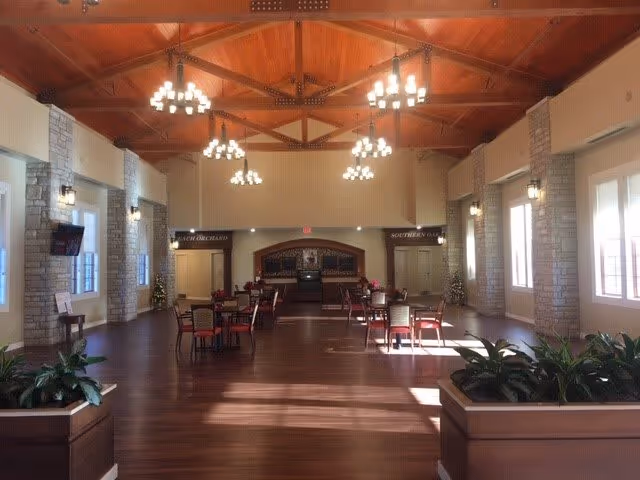 Spacious dining room with wooden floors, multiple tables and chairs arranged neatly, large windows letting in natural light, stone pillars along the walls, and chandeliers hanging from a high wooden ceiling.