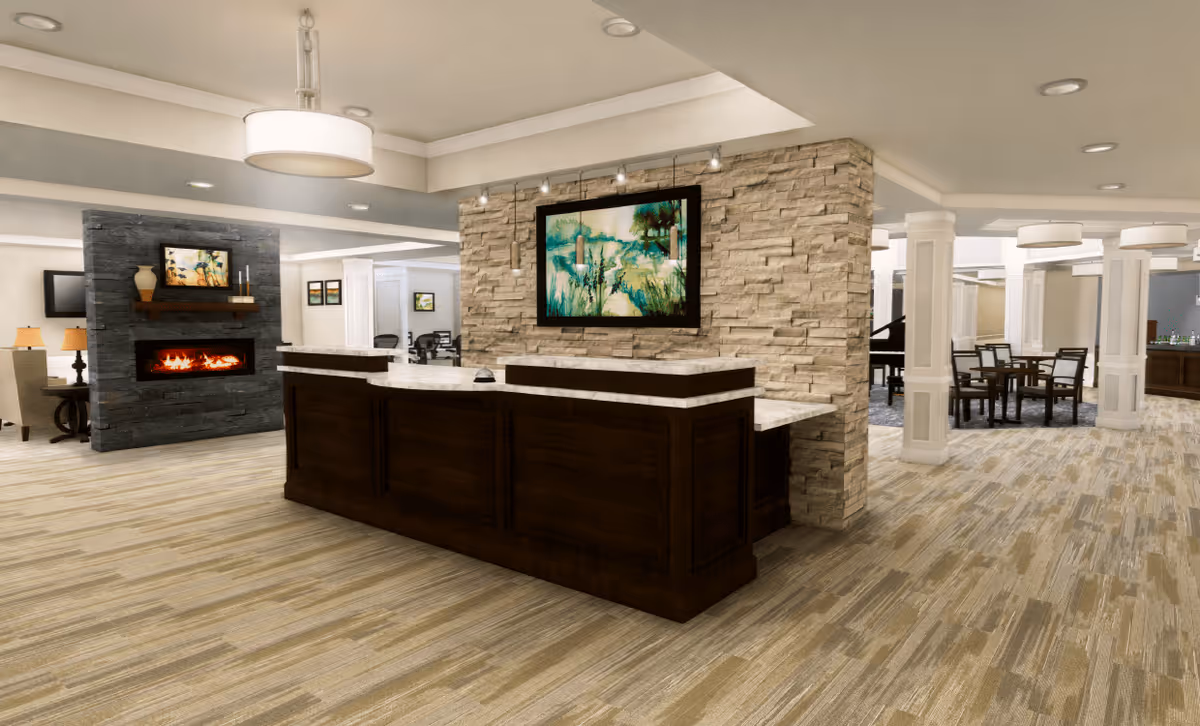 Interior view of a senior living facility lobby or common area featuring a dark wood reception desk with a marble countertop, a stone accent wall with a framed painting, a modern fireplace with a mantle and decorative items, and a seating area with tables and chairs in the background.