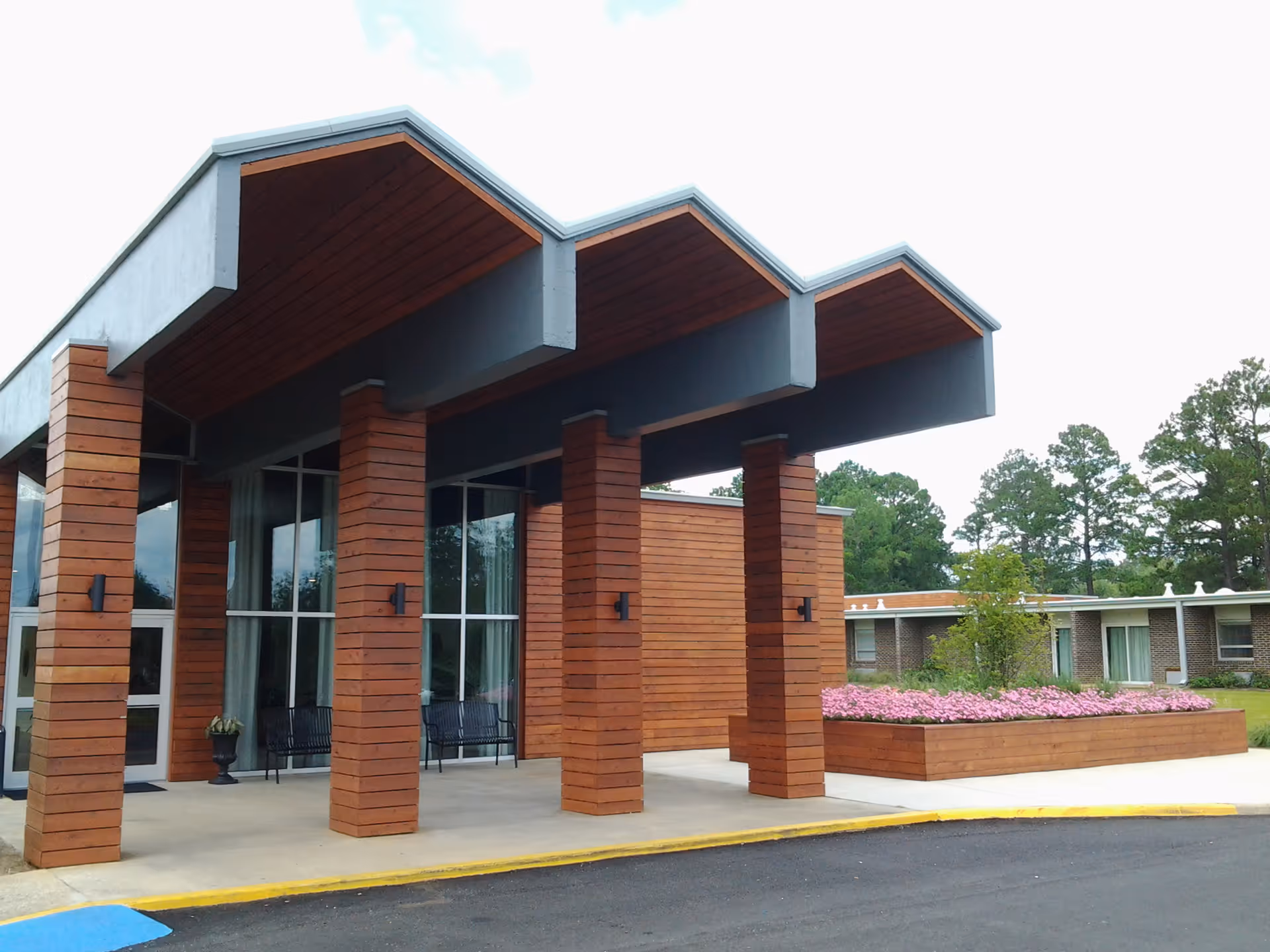Covered building entrance with wooden columns, large windows, and a raised flower bed in front of a low brick building and trees.