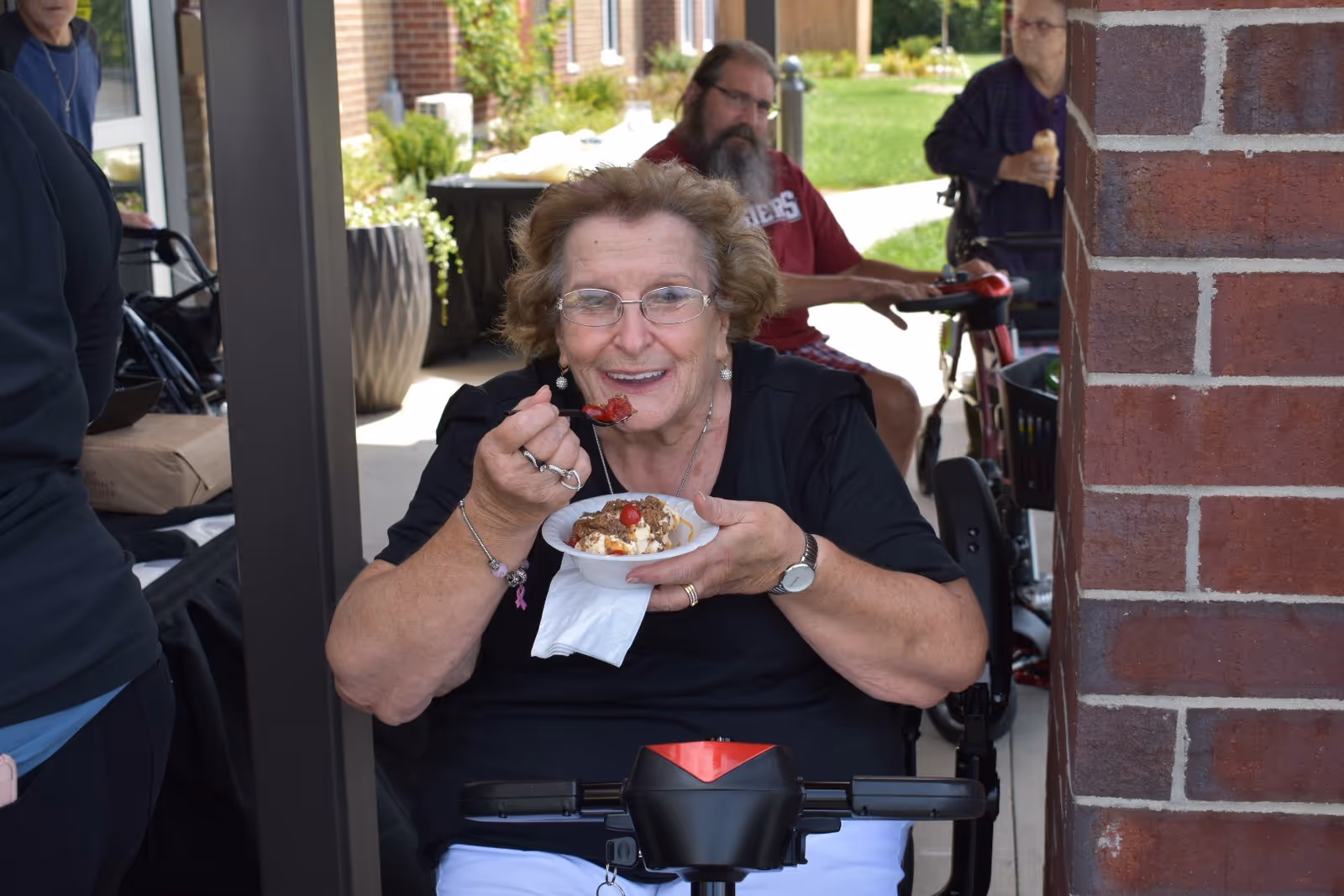An elderly woman sitting on a mobility scooter is smiling and eating a dessert from a small bowl with a spoon. Behind her, there are two other elderly individuals, one sitting on a mobility scooter and another standing, both enjoying ice cream cones. The setting appears to be an outdoor patio area with brick walls and greenery in the background.