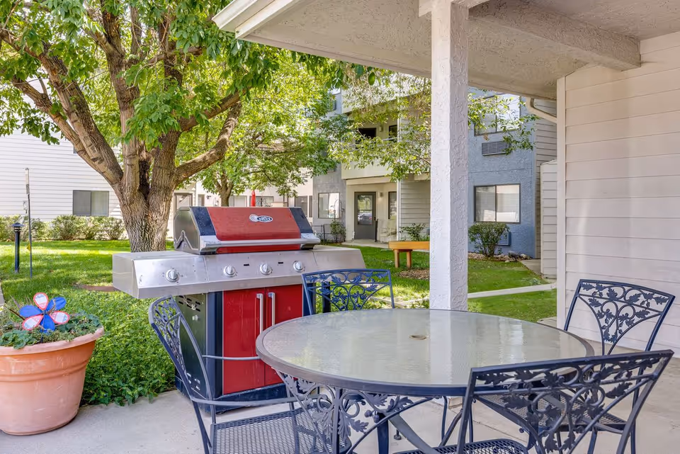 Outdoor patio area with a round glass-top table and four ornate metal chairs. A large stainless steel and red grill is positioned nearby. The patio is shaded by a roof and surrounded by green grass, trees, and residential buildings in the background. A large potted plant with a decorative pinwheel is also visible.
