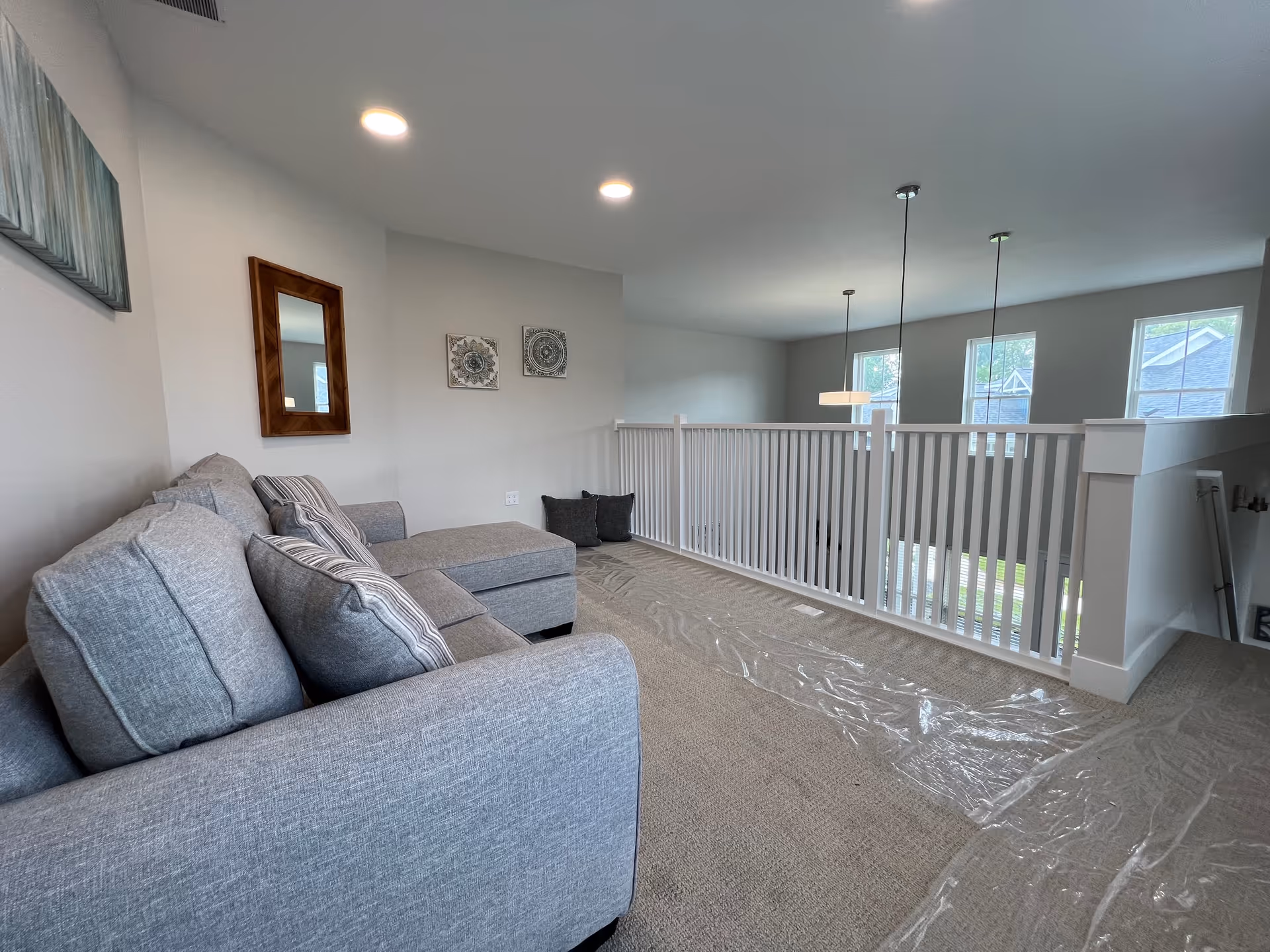 Bright upstairs loft living area with a gray sectional sofa, decorative wall art, and a white railing overlooking the lower level.