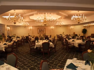 A spacious dining room with multiple round and rectangular tables covered with white tablecloths, green napkins, and floral centerpieces. The room features elegant chandeliers hanging from a high ceiling, patterned carpet flooring, and framed artwork on the walls.